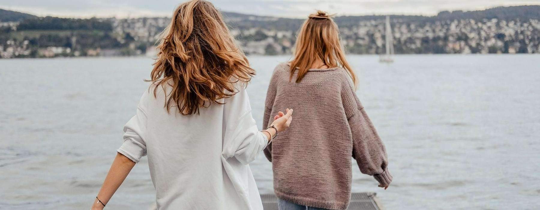 two women walk to the edge of a dock, in a body of water, looking toward the city on the other shore